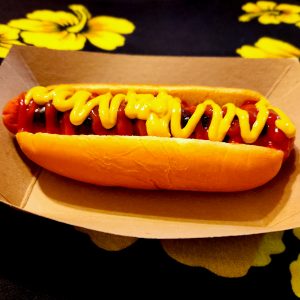 Hot dog with mustard and ketchup for toppings on a brown paper food tray. Background is black with yellow flowers.