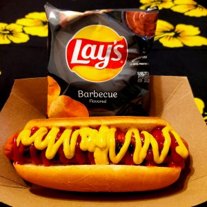 Hot dog with mustard and ketchup for toppings on a brown paper food tray. A bag of Lays barbecue chips sits on the brown paper food tray next to the hot dog. Background is black with yellow flowers.